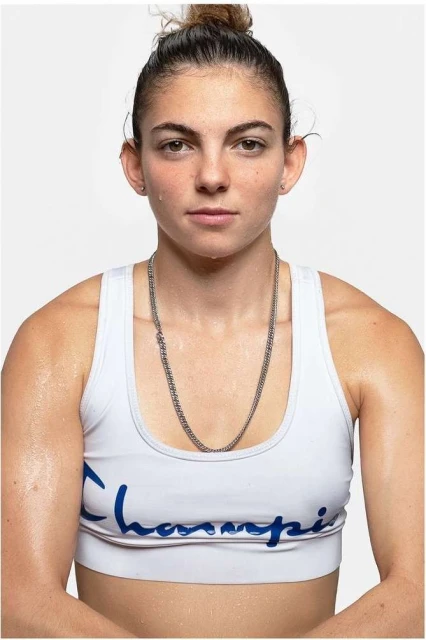 Portrait of a sweaty woman wearing a white sports bra and silver necklace against a white backdrop in an eye-level studio shot.