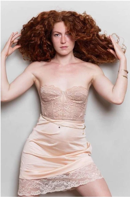 A woman with curly red hair poses in a lace bustier and slip dress against a neutral background, looking directly at the camera.