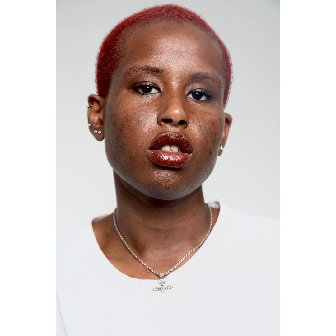 Close-up portrait of a young adult woman with short, dyed red hair wearing a white t-shirt and a silver necklace against a light grey backdrop.