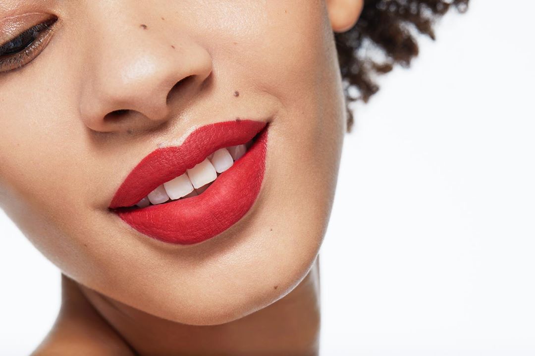 Close-up portrait of an adult woman smiling with bright red lipstick, showcasing her teeth and radiant skin against a white background.