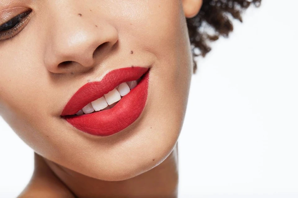 Close-up portrait of an adult woman smiling with bright red lipstick, showcasing her teeth and radiant skin against a white background.
