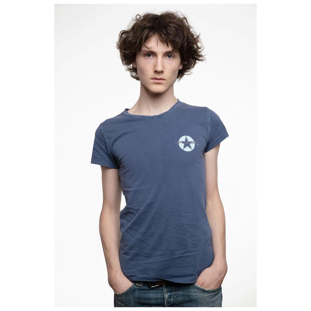 Portrait of a young adult male with curly brown hair, wearing a blue t-shirt with a star logo, set against a clean white backdrop.