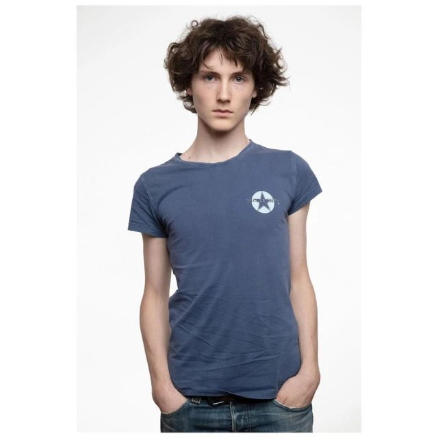 Portrait of a young adult male with curly brown hair, wearing a blue t-shirt with a star logo, set against a clean white backdrop.