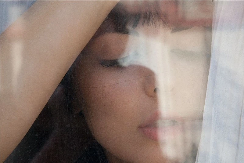 Close-up portrait of a young adult woman with closed eyes, her face partially visible through textured glass, soft lighting, muted color palette.