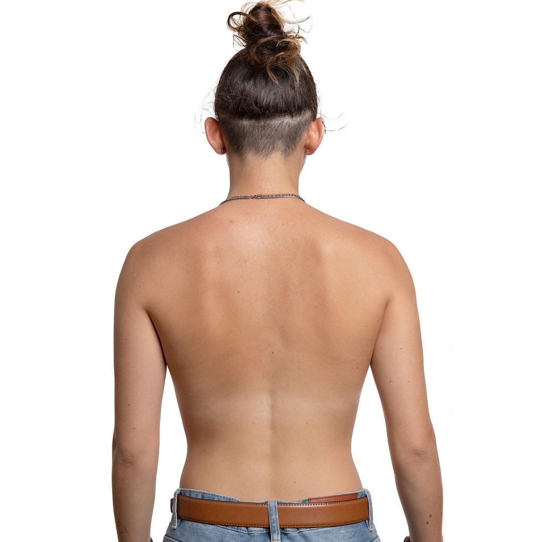Woman's bare back, turned away from camera, hair in messy bun. Tan skin with tan line on her back. Wearing blue denim jeans and a brown belt.