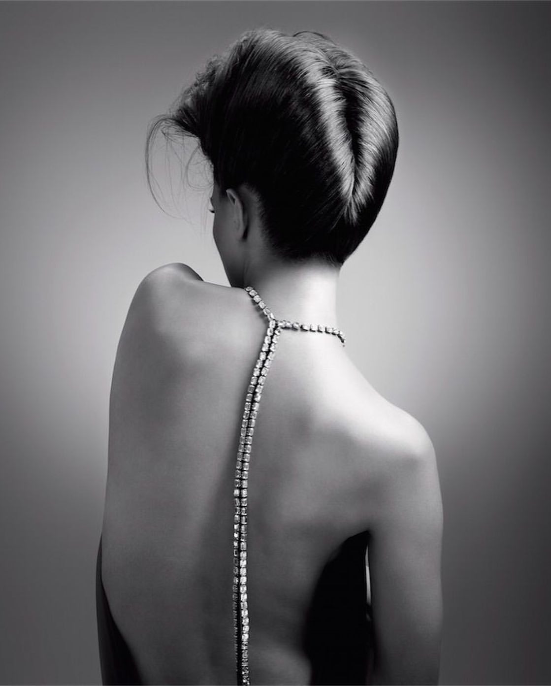 Model seen from behind wearing an elaborate updo hairstyle and a long diamond necklace down her bare back against a gradient backdrop.