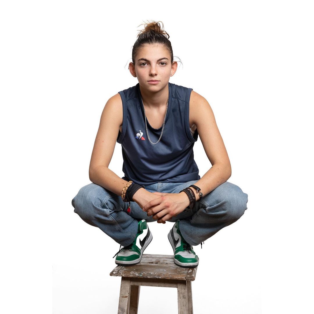 Young woman in a dark blue tank top and light wash jeans squatting on a wooden stool against a white background