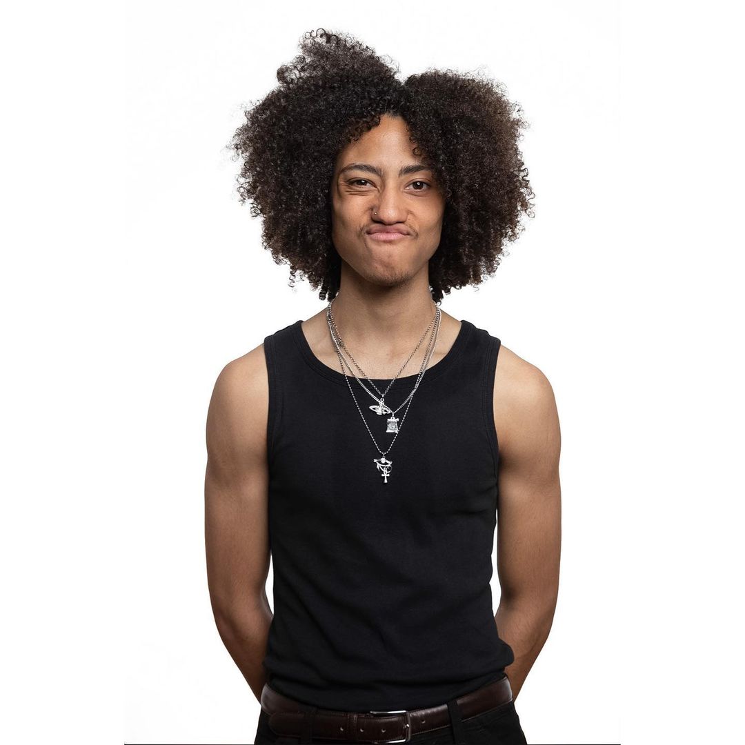 Young adult with afro, wearing a black tank top and necklaces, makes a silly face in a studio portrait on a white background.