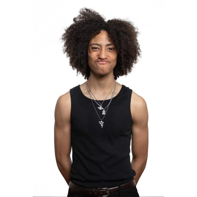 Young adult with afro, wearing a black tank top and necklaces, makes a silly face in a studio portrait on a white background.
