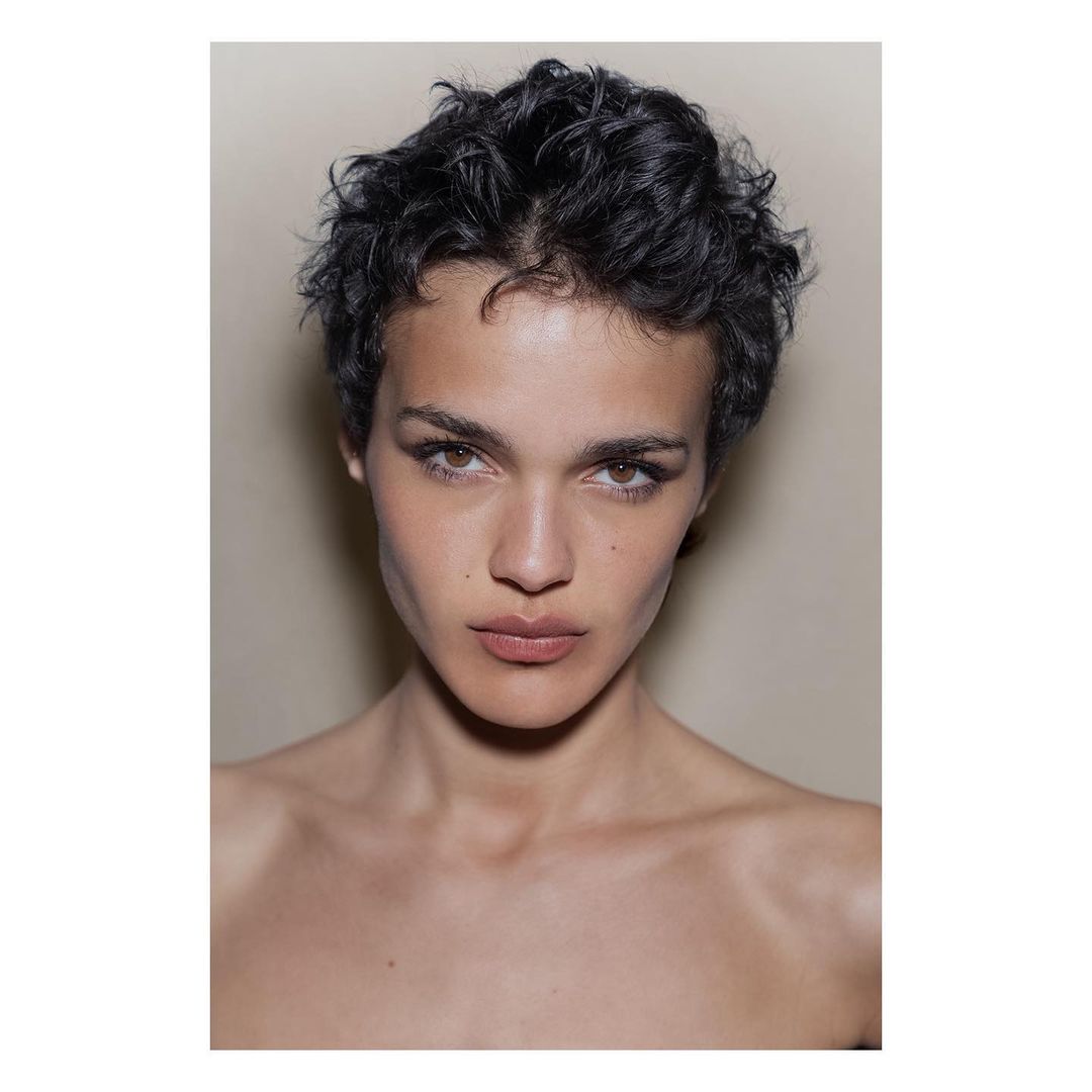 Close-up portrait of a young adult woman with short curly black hair and light brown eyes, set against a neutral beige background.
