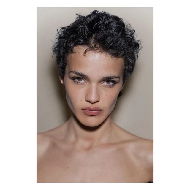 Close-up portrait of a young adult woman with short curly black hair and light brown eyes, set against a neutral beige background.