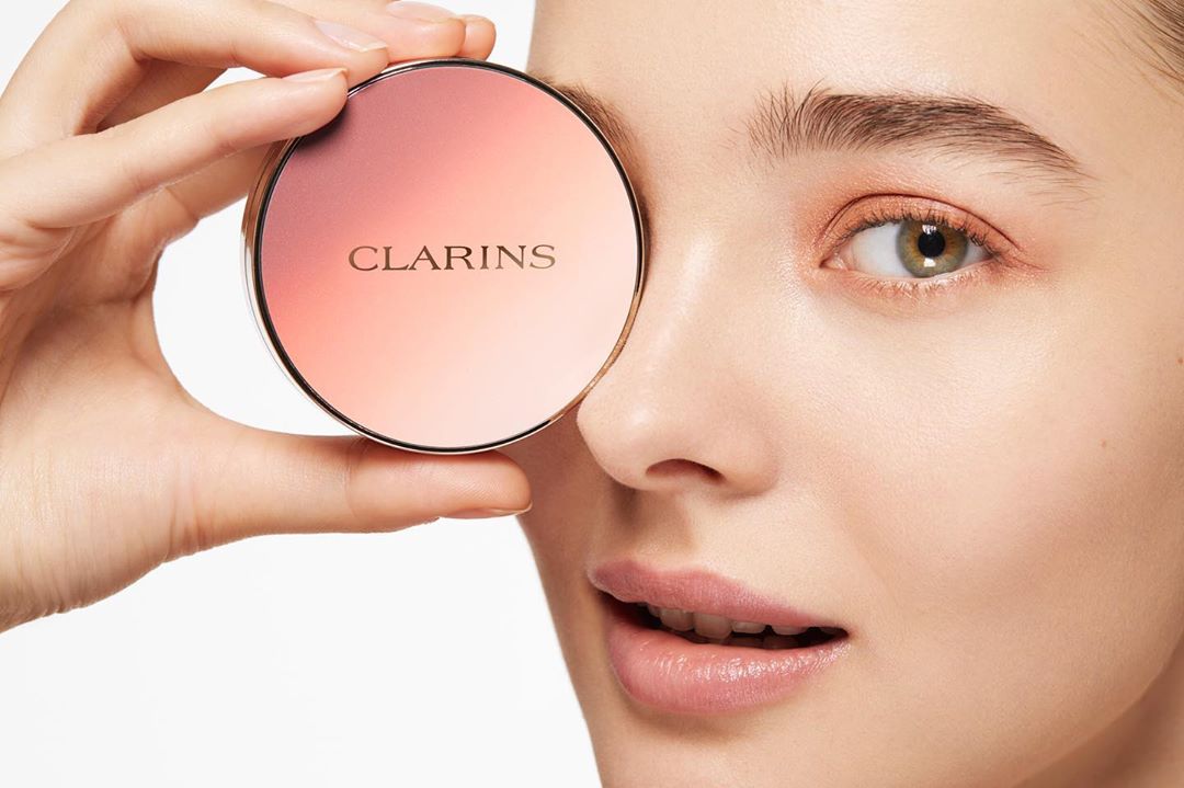 Close-up of a woman holding a Clarins compact. The make-up product is featured alongside the woman's face, emphasizing beauty and skincare.