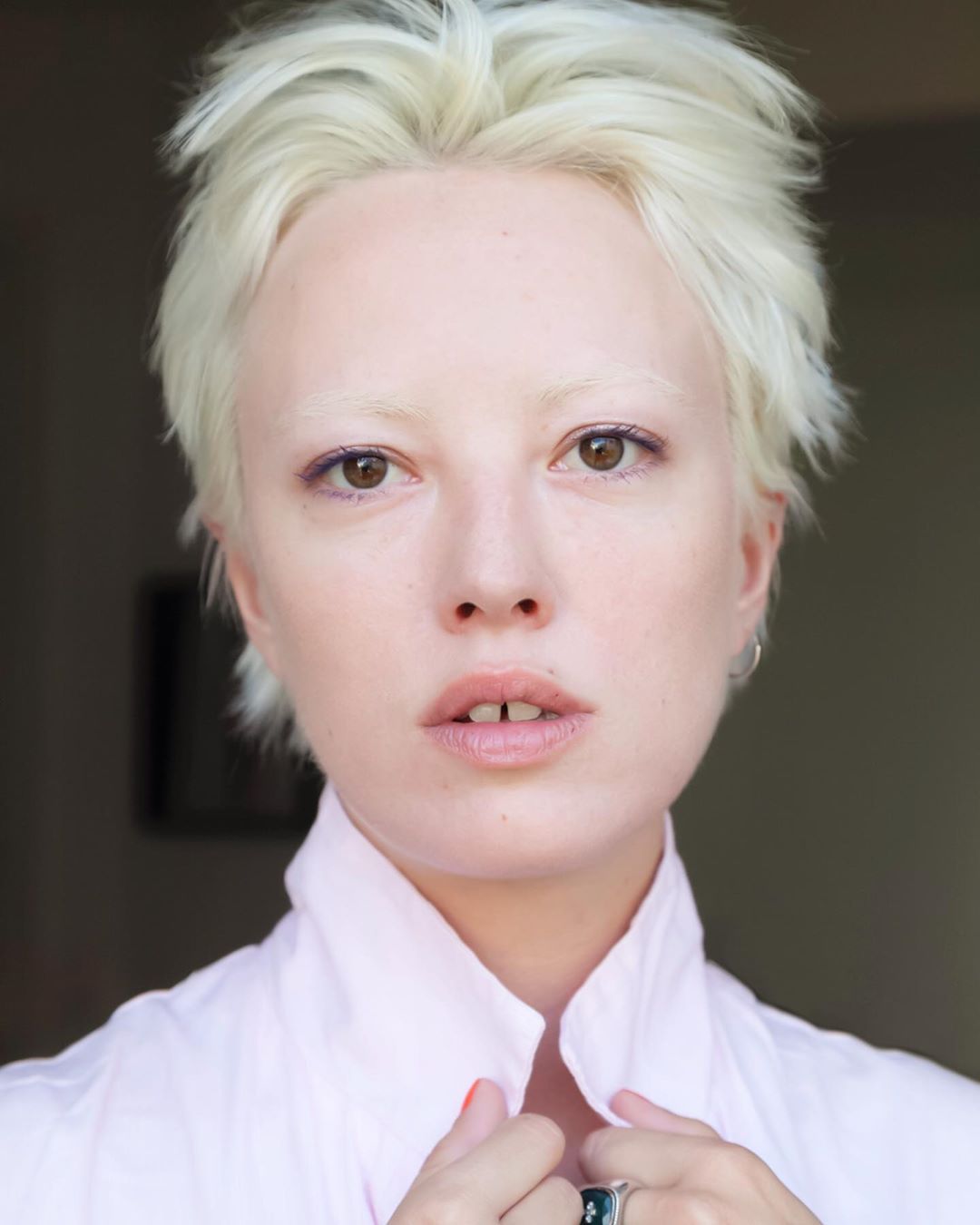 Close-up portrait of a woman with short white hair, wearing a pink collared shirt and ring. She has light skin and is indoors.