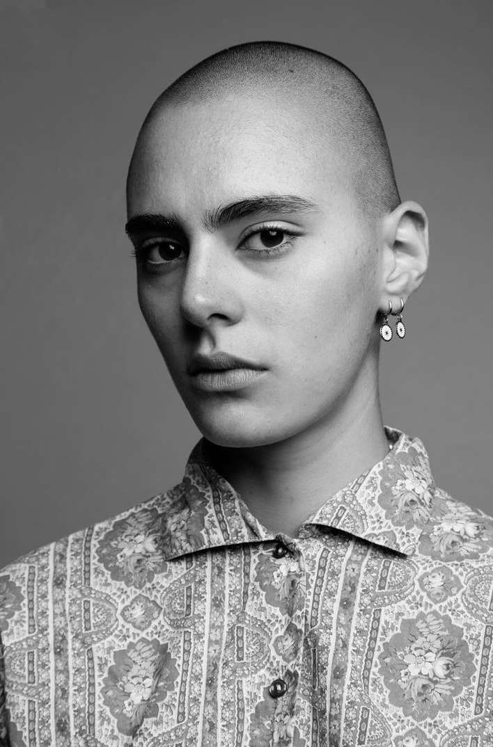 Striking black and white portrait of a bald woman in a patterned shirt, gazing directly at the camera with earrings. Simple grey backdrop.