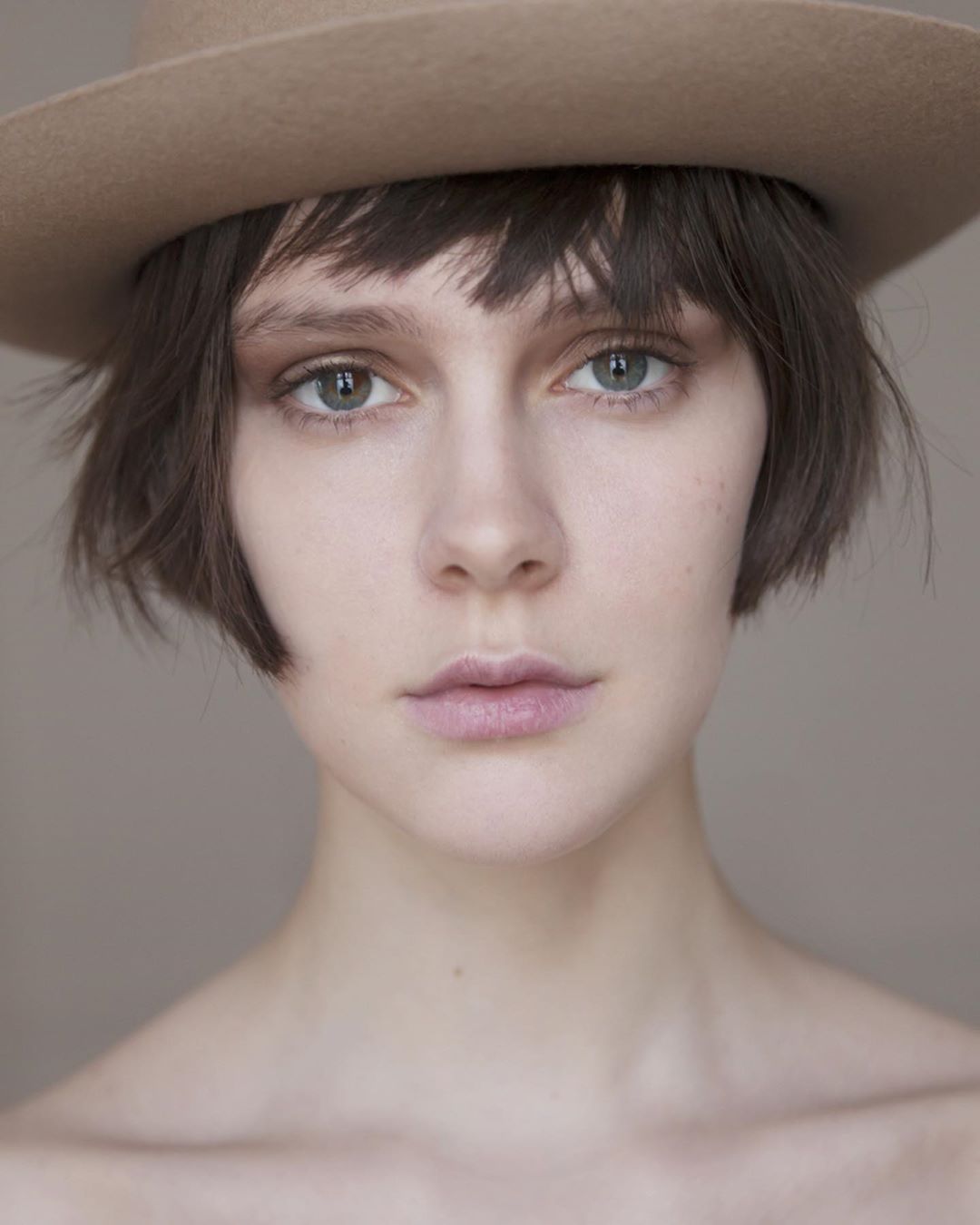 Close-up portrait of a young adult woman with short brown hair wearing a tan hat against a neutral background in soft, diffused lighting.