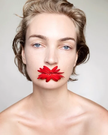 Close-up portrait of a woman with short blonde hair holding a vibrant red flower between her lips against a neutral background.