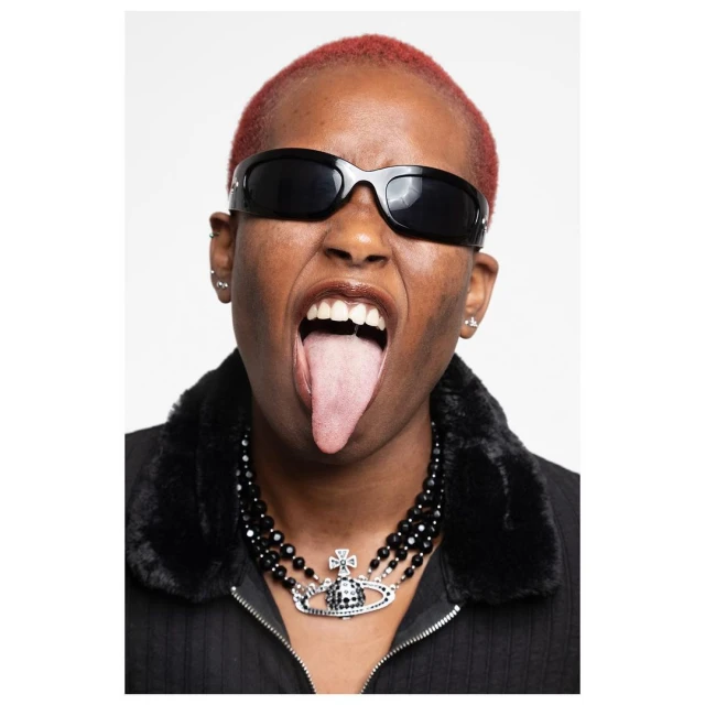 Eye-level portrait of a person with red hair, wearing sunglasses and a black necklace, sticking their tongue out against a white background.