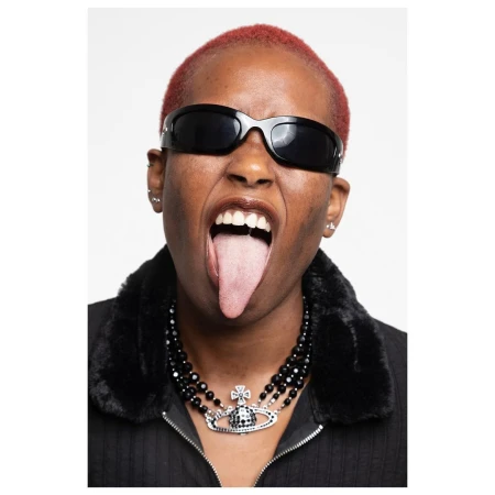 Eye-level portrait of a person with red hair, wearing sunglasses and a black necklace, sticking their tongue out against a white background.