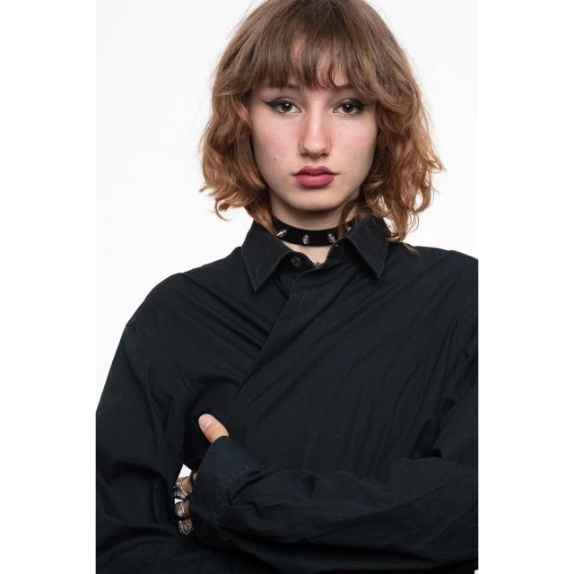 Young woman with wavy brown hair and dark eyes wearing a black button-down shirt and studded choker against a white backdrop.