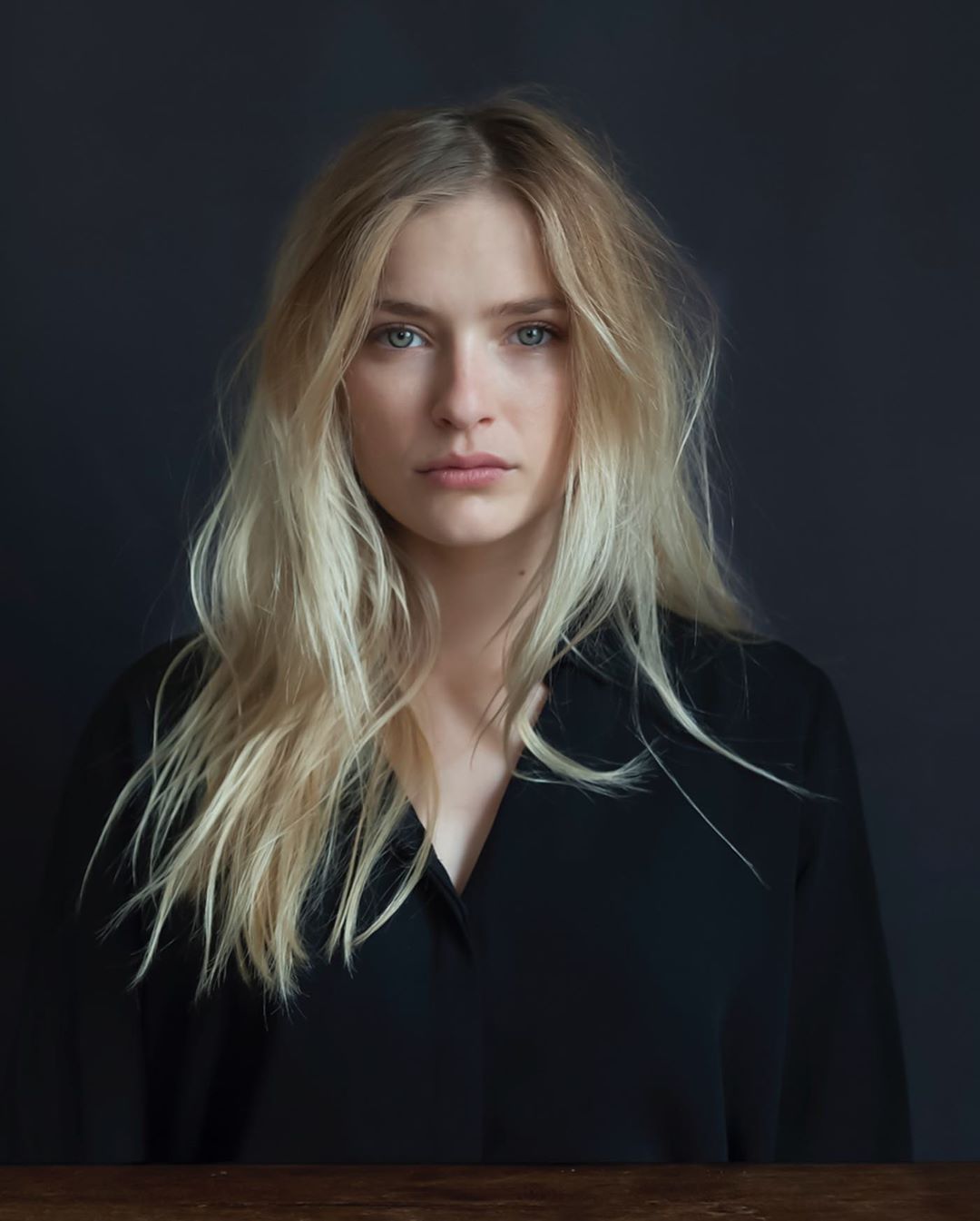 Portrait of a blond woman with light eyes wearing a black shirt against a dark backdrop and a wooden table.