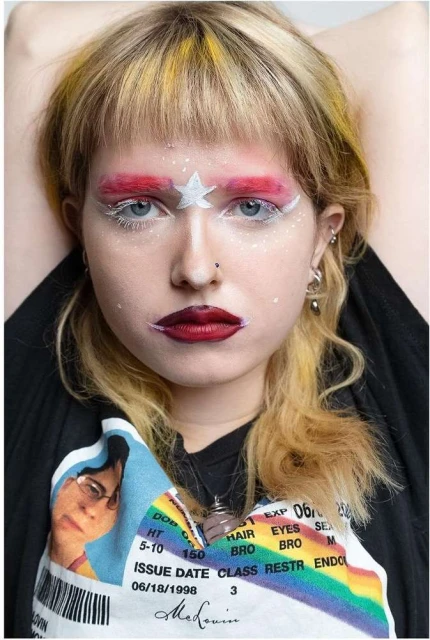 Close-up portrait of a young woman with unique makeup and a T-shirt featuring an image and text, posing with her hands behind her head.