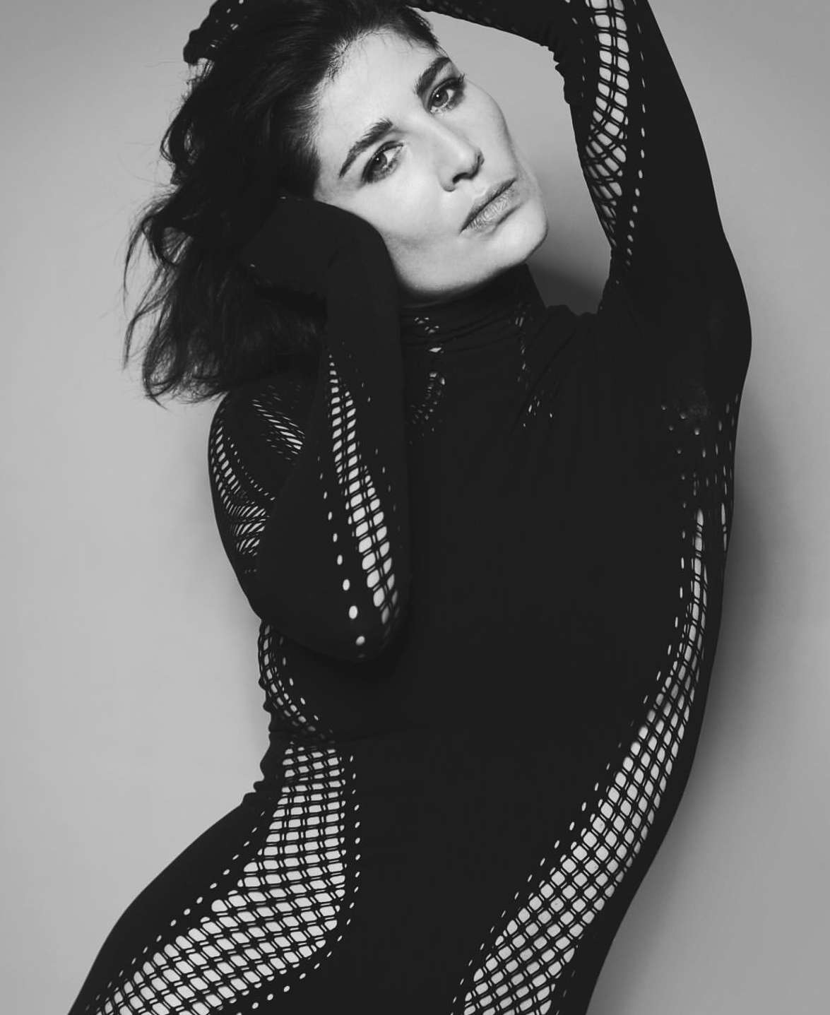 Monochrome portrait of an adult woman in a black mesh bodysuit, posing with arms raised against a plain grey backdrop in a studio setting.