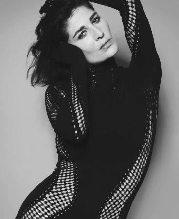Monochrome portrait of an adult woman in a black mesh bodysuit, posing with arms raised against a plain grey backdrop in a studio setting.