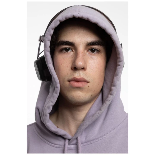 Portrait of a young adult wearing a lilac hooded sweatshirt and black headphones against a plain white studio backdrop.