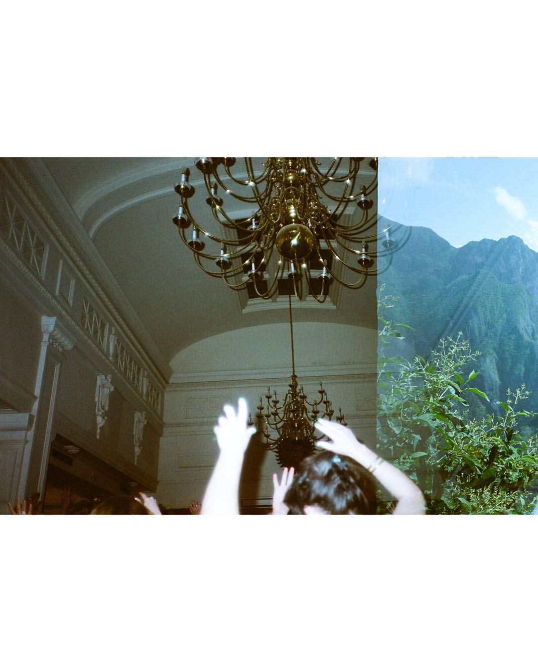 Indoor scene with a chandelier and people contrasted with an outdoor mountain landscape with lush vegetation under a bright blue sky.