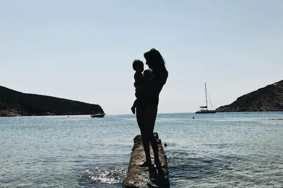 Silhouette of an adult female holding a baby on a concrete pier overlooking the sea, with islands and boats in the distance.