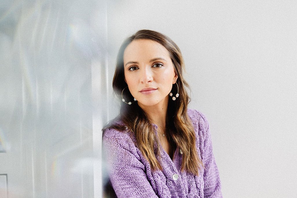 Portrait of an adult woman with long brown hair wearing a lilac knit sweater and hoop earrings with pearls, posing indoors against white.