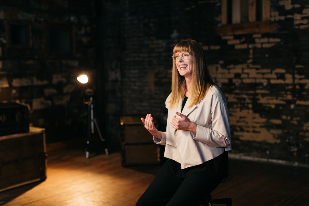 Smiling woman with blonde hair and glasses in a studio. Wearing a light jacket, she gestures against a dark brick background with soft lighting.