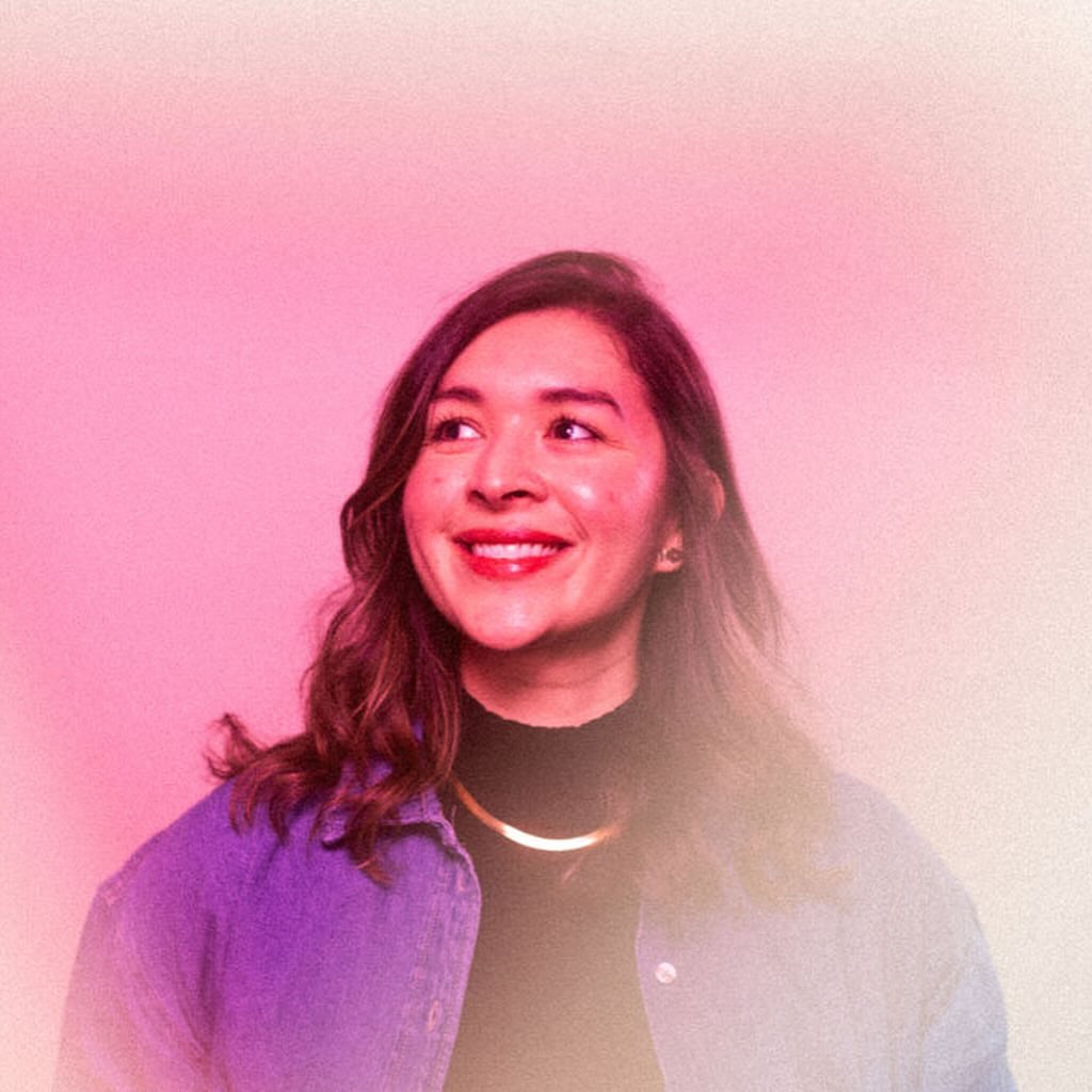 Portrait of a smiling woman in her 20s wearing a blue denim jacket over a black turtleneck against a pink gradient background.