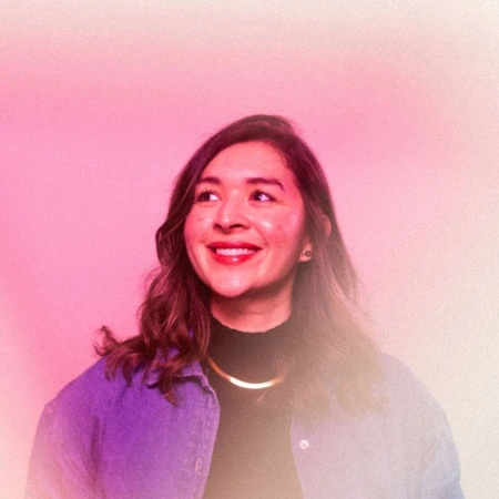 Portrait of a smiling woman in her 20s wearing a blue denim jacket over a black turtleneck against a pink gradient background.