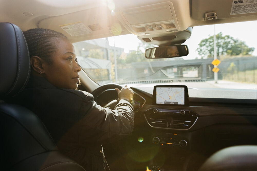 Black woman driving car with navigation system in bright sunlight. Interior view of car dashboard, steering wheel, and road visible.