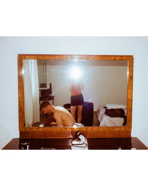Reflection in a vanity mirror shows a shirtless adult male and a female taking the picture in a bedroom with a bed and luggage.