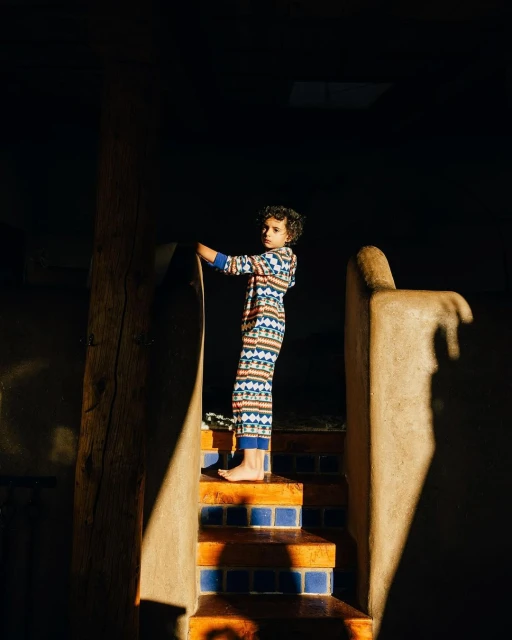 Child in patterned pajamas stands on tiled stairs indoors with dark background. Warm lighting highlights the textures and colors.