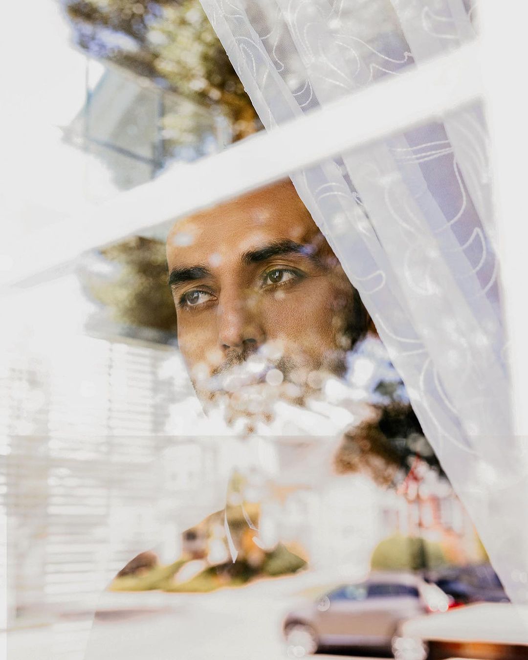 Adult male gazes pensively out a window with lace curtains, reflecting an outdoor scene with cars and foliage in soft focus.