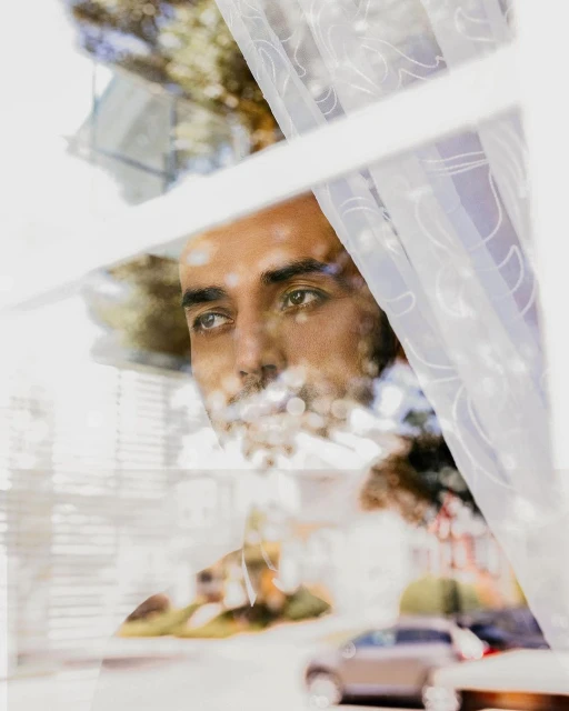 Adult male gazes pensively out a window with lace curtains, reflecting an outdoor scene with cars and foliage in soft focus.