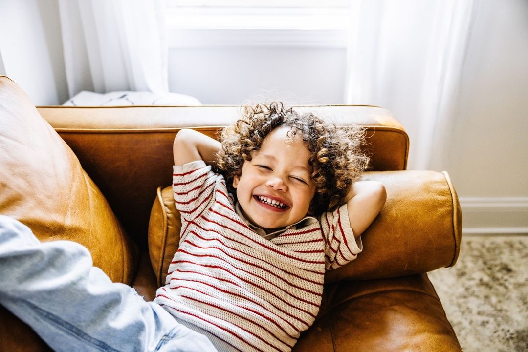 Happy curly-haired child laughs on a brown leather couch in bright light with their hands behind their head; casual indoor portrait.