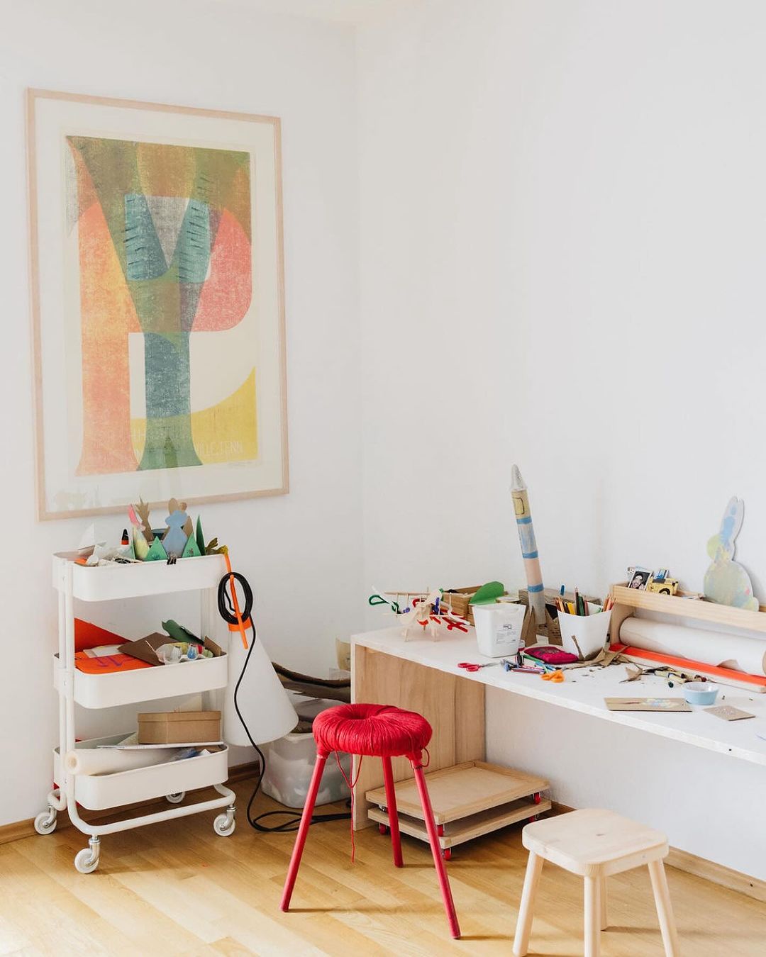 Bright playroom interior with art, craft table, and storage cart, featuring a red stool and light wood flooring, capturing a creative space.