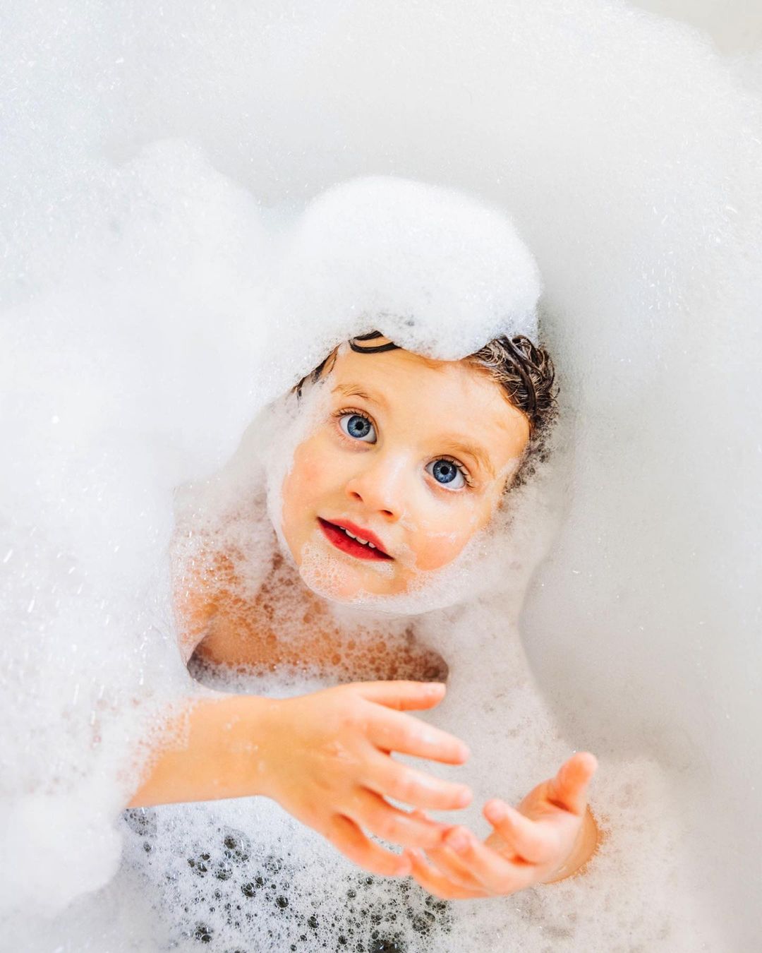 Child in bathtub covered in white soap bubbles. Child has wet hair and blue eyes, looking up. Cleanliness, health, and hygiene.