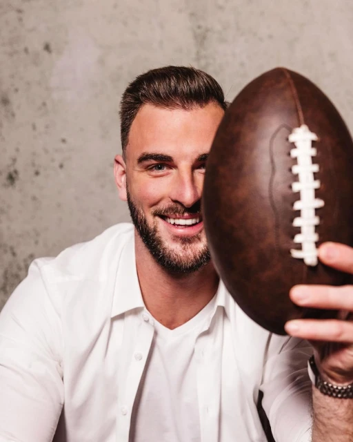 Adult male smiles broadly, holding a brown leather American football in a studio setting. Focus on face and ball.