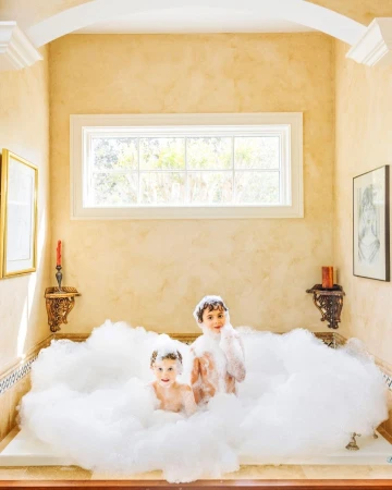 Two young children covered in bubbles play together inside a vintage bathtub in a brightly lit bathroom with soft yellow walls.