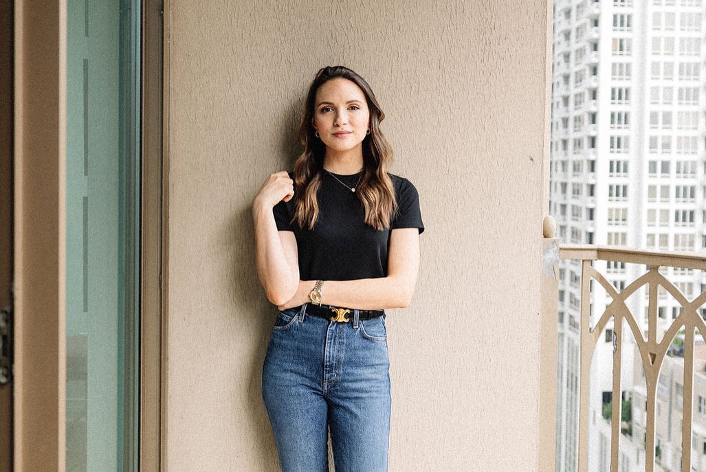 Portrait of a woman in a black t-shirt and jeans posing on a balcony, with high-rise buildings visible in the background.