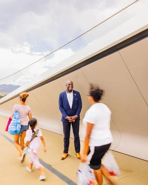 A well-dressed Black man in a suit stands smiling on a modern bridge with blurred people walking by on a bright day with cloudy skies.