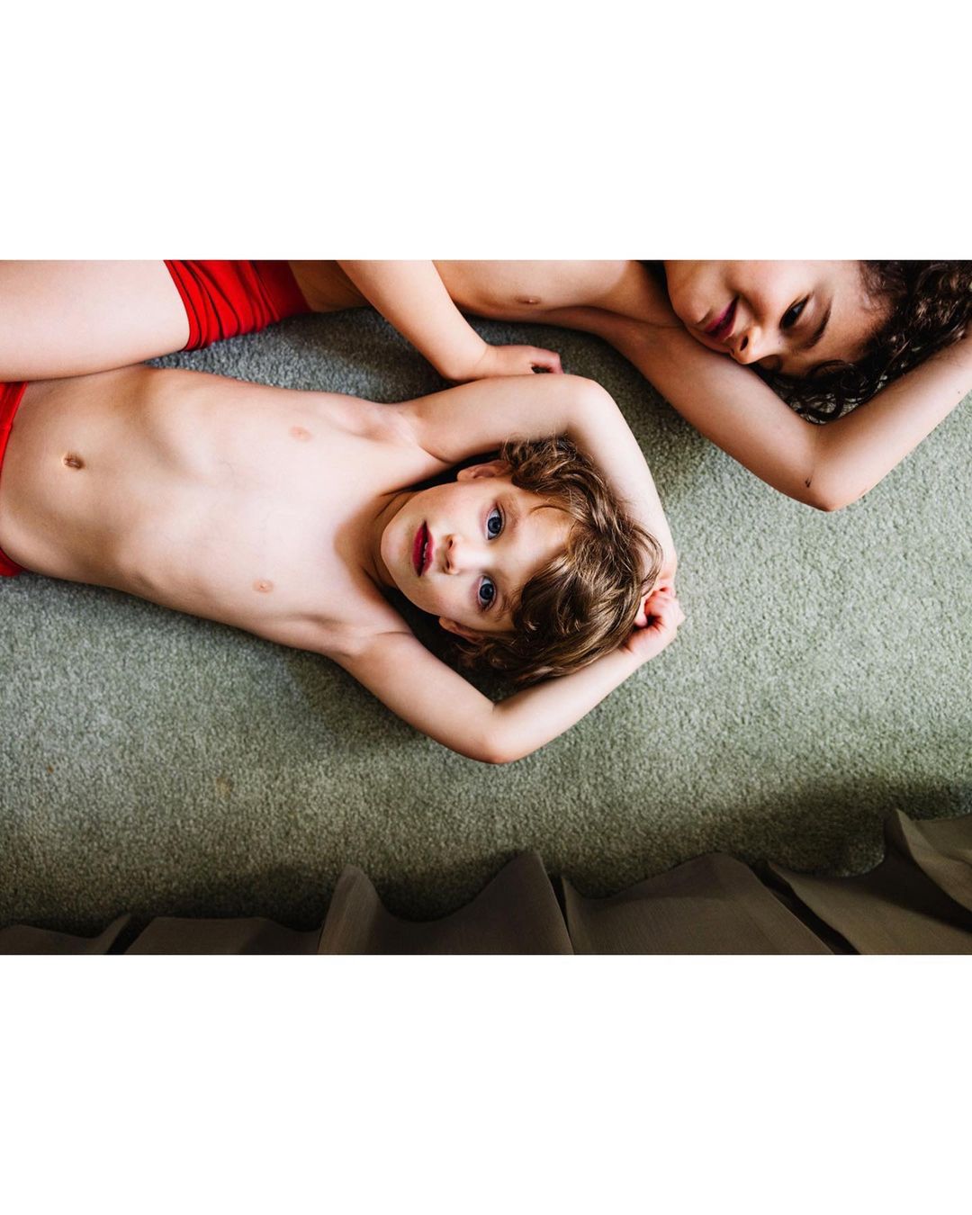 Two shirtless children, one boy with blue eyes and one girl with curly hair, lie on a green carpet indoors, in an eye-level, medium shot.