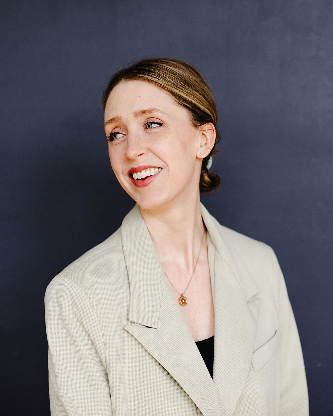 Smiling woman in a sand-colored blazer against a dark backdrop, captured in a studio portrait with soft lighting and natural makeup.
