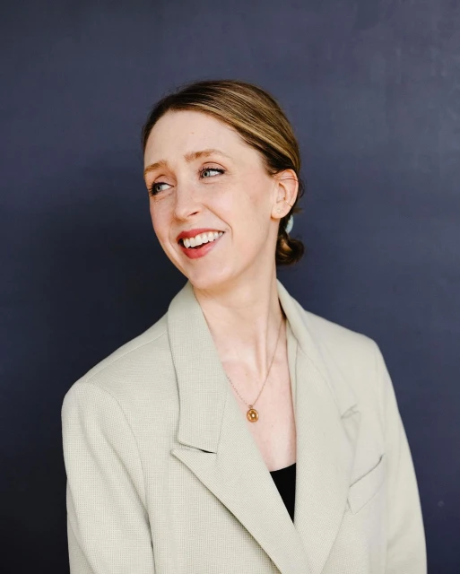 Smiling woman in a sand-colored blazer against a dark backdrop, captured in a studio portrait with soft lighting and natural makeup.