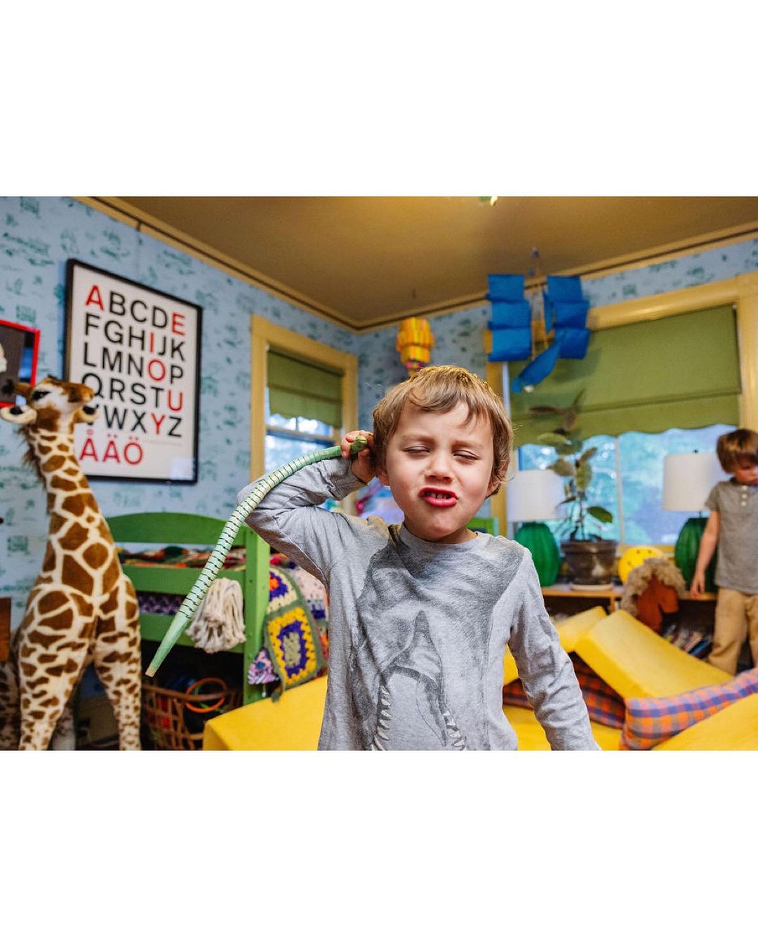 A young boy makes a face indoors while holding a green hose up to his ear, with another child and a toy giraffe visible in the background.